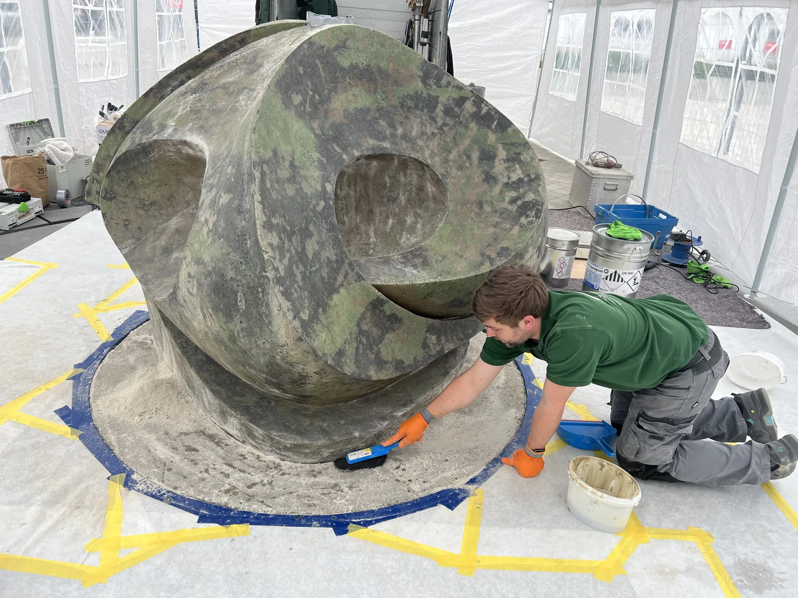 Ein Mann arbeitet unter einer Zeltplane an der dunklen Wellen-Skulptur des Brunnens