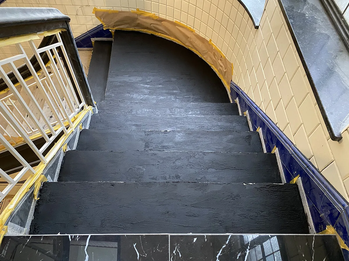 Frisch mit schwarzem Sichtbeton beschichtete Treppenstufen, daneben gelbe Wandfliesen mit blauem Band und abgeklebte Kanten